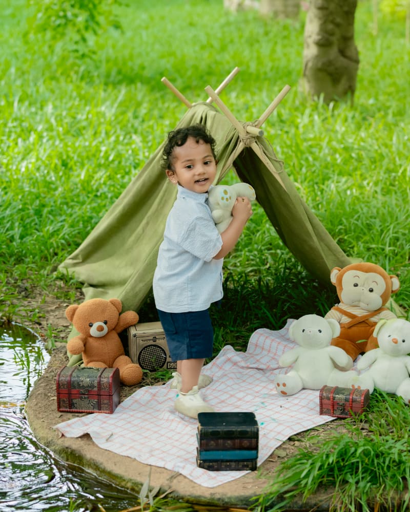 A little boy standing by a green tent with teddies on a grassy lakeside