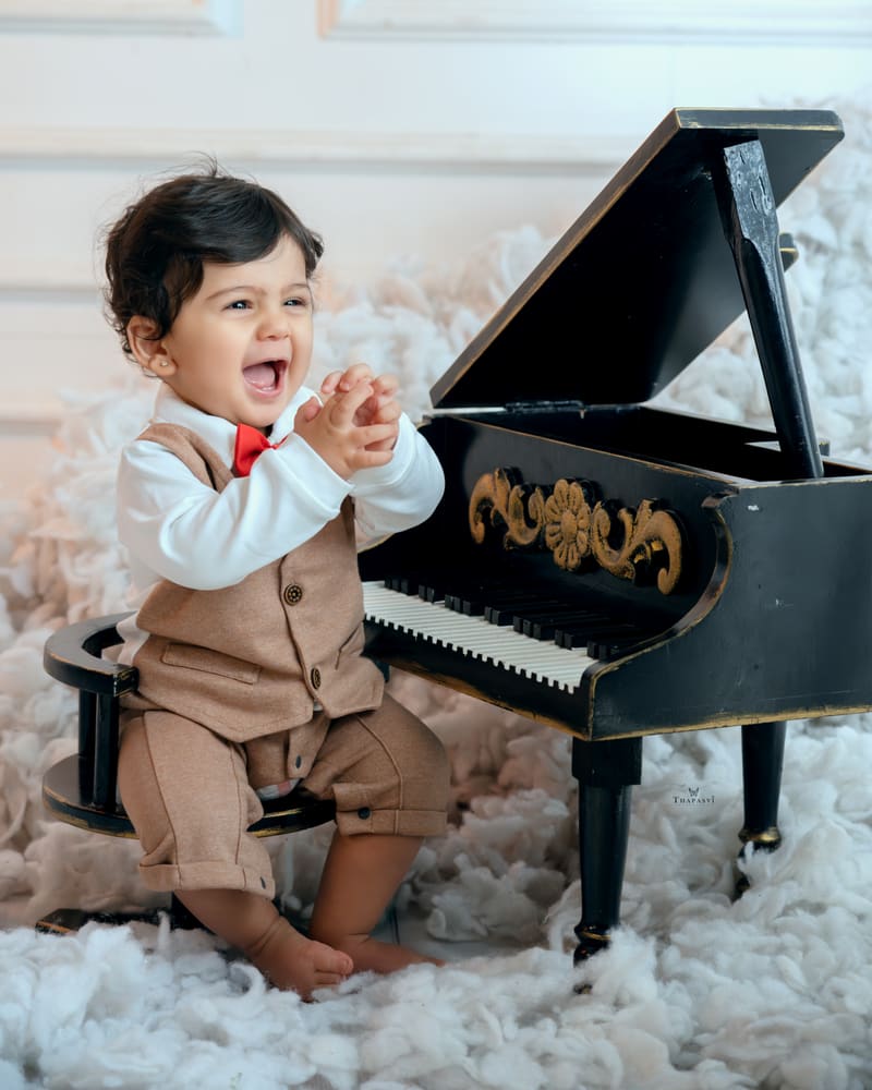 A laughing little boy in a waistcoat and bow tie at a toy grand piano on a white set