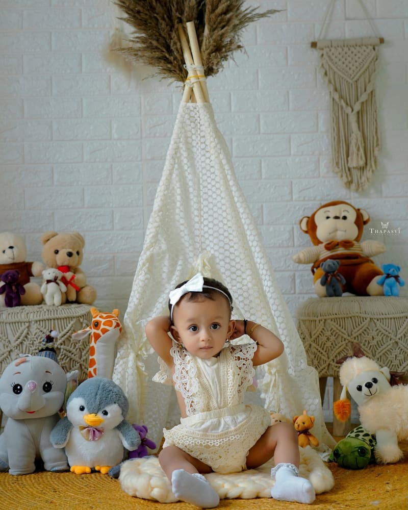 A baby girl in a cream romper in front of a lace tipi and soft toys