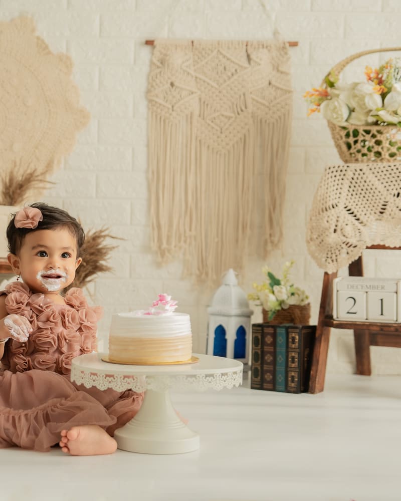 A baby girl in a blush dress beside a cake on a macramé set