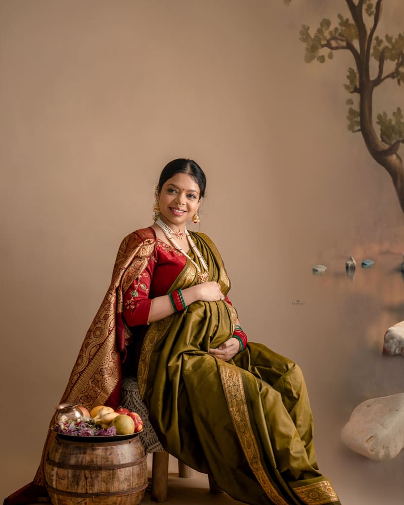 A mother-to-be in an olive silk saree, seated beside a wooden barrel