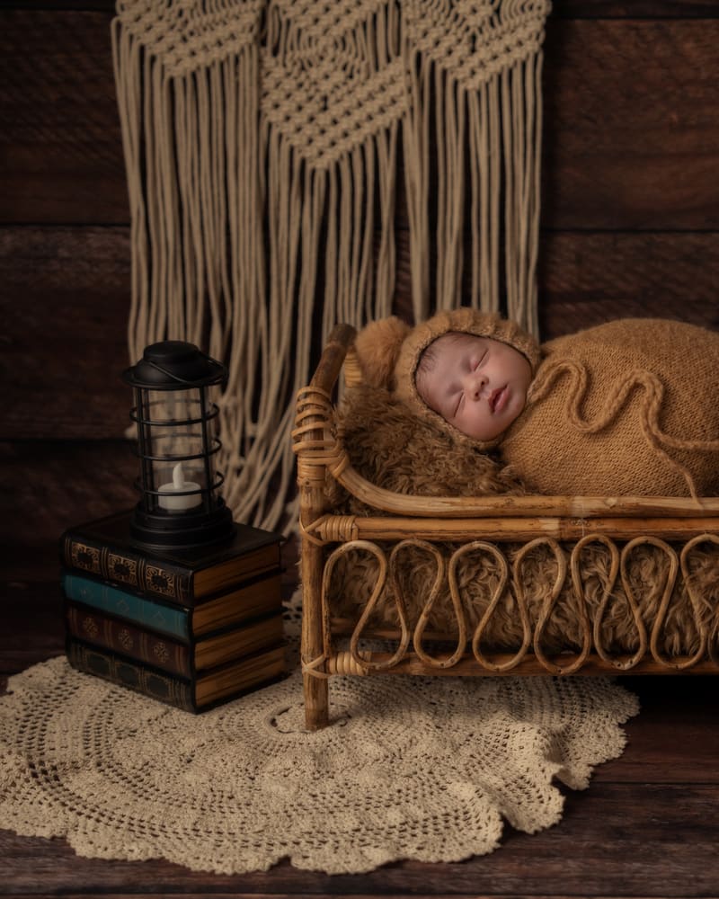 A newborn asleep in a rattan bed on a dark, rustic set with macramé