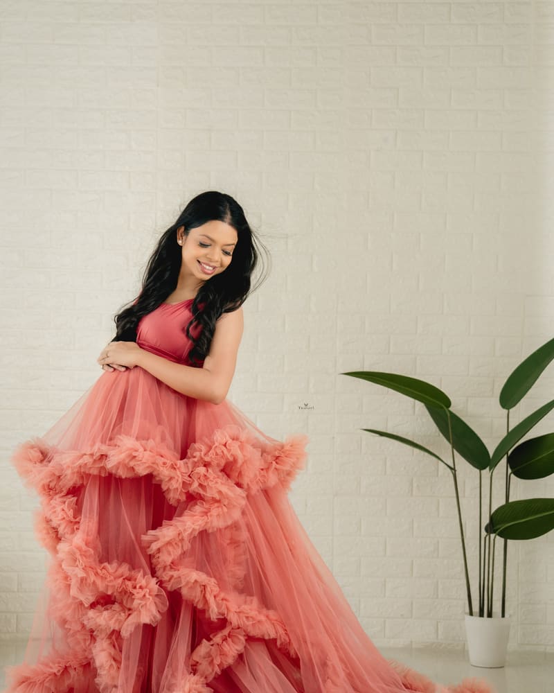 A mother-to-be in a coral ruffled gown against a white wall