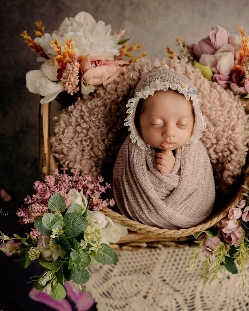 A newborn in a dusty-pink bonnet and wrap among muted flowers