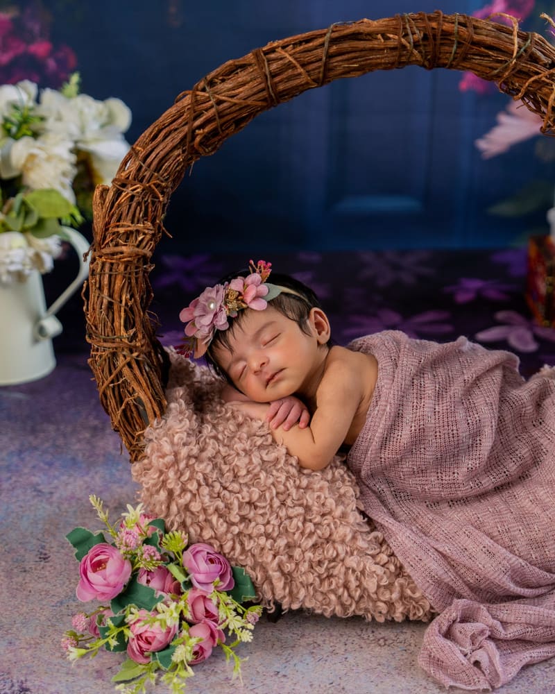 A newborn asleep in a woven basket with pink flowers and a mauve throw