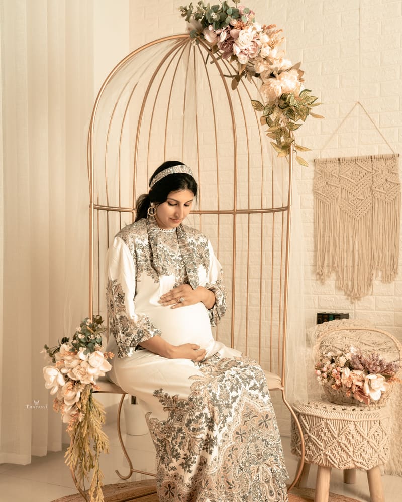 A mother-to-be in an ivory gown seated in a gold birdcage chair on a boho cream set