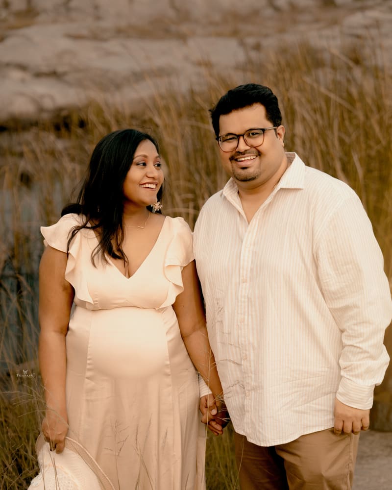 An expecting couple in cream and white, standing in golden grass outdoors